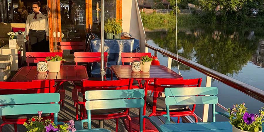  Vibrant open sun deck on the barge, with red and turquoise chairs, and flowers on each table. 